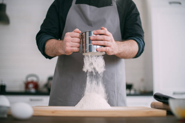Egy férfi séf tésztát készít otthon a konyhában. Fiatal fickó kötényben a konyhában az asztalnál, hozzávalókkal a tésztakészítéshez. - Fotó, kép