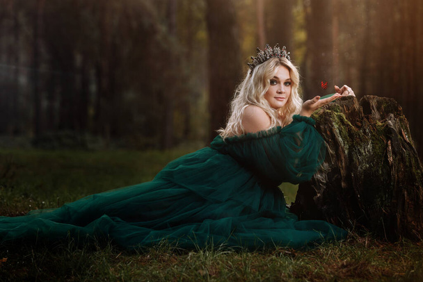 Eine schöne blonde junge Frau in einem langen grünen Kleid und einem Diadem auf dem Kopf im Wald. Mädchen, das neben dem alten Baumstumpf mit einem Schmetterling auf der Hand sitzt. Sonnenblendung. Fantasie. Märchen. - Foto, Bild