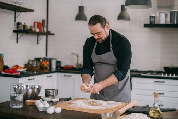 Un chef masculin prépare de la pâte à la maison dans la cuisine. Jeune homme dans un tablier dans sa cuisine à la maison à la table avec des ingrédients pour faire de la pâte - Photo, image