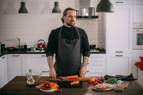 Erkek bir aşçı mutfakta evde sebze hazırlar. Evdeki mutfakta önlüklü genç adam sebze güveci ya da salata yapmak için malzemelerle birlikte. - Fotoğraf, Görsel