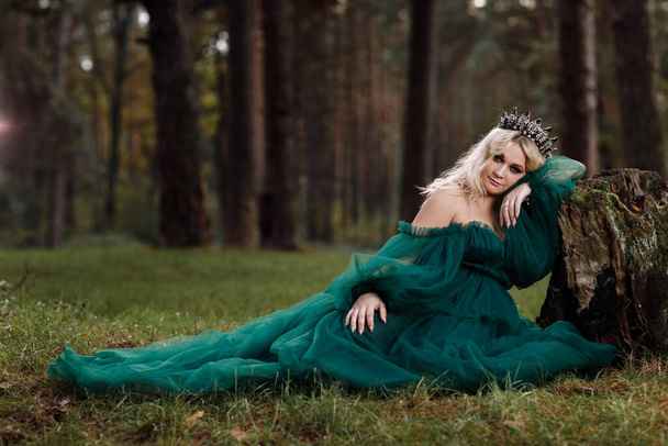 Eine schöne blonde junge Frau in einem langen grünen Kleid und einem Diadem auf dem Kopf im Wald. Mädchen, das neben dem alten Baumstumpf sitzt. Sonnenblendung. Fantasie. Märchen. - Foto, Bild