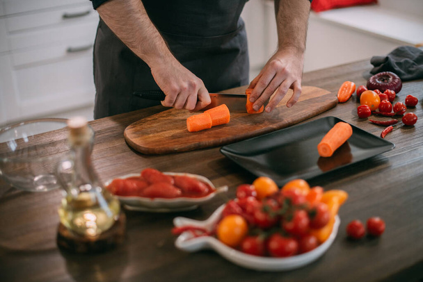 Ένας αρσενικός μάγειρας ετοιμάζει λαχανικά στο σπίτι στην κουζίνα. Κοντινό πλάνο των χεριών με ένα μαχαίρι και λαχανικά. Νεαρός με ποδιά στην κουζίνα του σπιτιού στο τραπέζι με υλικά για βραστό λαχανικών ή σαλάτα - Φωτογραφία, εικόνα