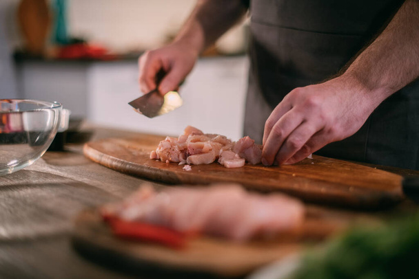 Um chef masculino prepara carne fresca em casa na cozinha. Feche as mãos com uma faca e um pedaço de carne. Jovem em um avental na cozinha na casa à mesa com ingredientes para cozinhar um prato de carne - Foto, Imagem