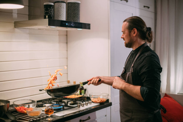 Miespuolinen kokki kokkaa keittiön uunissa. Nuori mies esiliinassa keittiössä kaasuliedellä paistaa ruokaa wokissa. - Valokuva, kuva