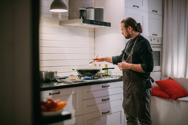 Un cuisinier masculin cuisine à la cuisinière à la maison dans la cuisine. Un jeune homme dans un tablier dans la cuisine à la cuisinière à gaz frit de la nourriture dans un wok - Photo, image
