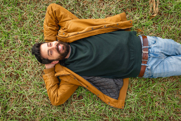 Молодой кавказский мужчина улыбается, лежа на траве, человек носит джинсы, зеленый свитер и коричневую куртку. - Фото, изображение