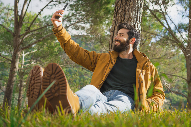 Young Caucasian man sitting in a Mediterranean forest leaning on a pine tree, taking a selfie for his social networks. - Foto, Bild
