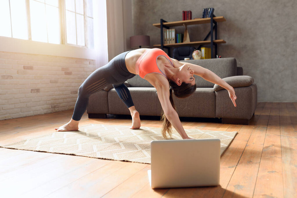 A mulher jovem segue com um computador portátil uns exercícios de ginásio. Ela está em casa devido à quarentena do coronavírus codiv-19. - Foto, Imagem