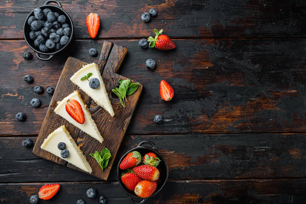Cheesecake with blueberrie and strawberries, on old dark  wooden table background, top view flat lay with copy space for text - Фото, изображение