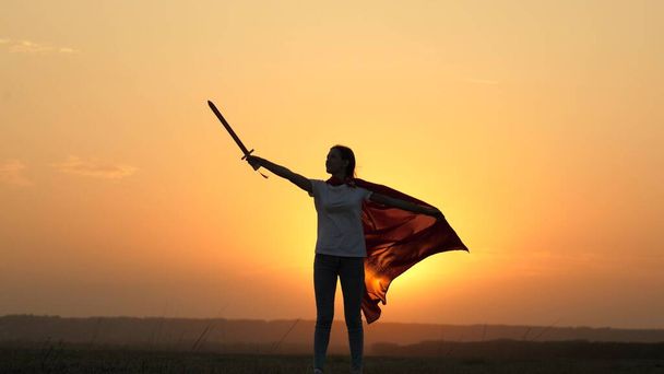 Girl fights with a toy sword. A free girl in a red cloak with a sword in her hand plays a medieval knight in the sun. Young girl plays superheroes. Child plays Spartan In. Happy childhood concept. - Photo, Image