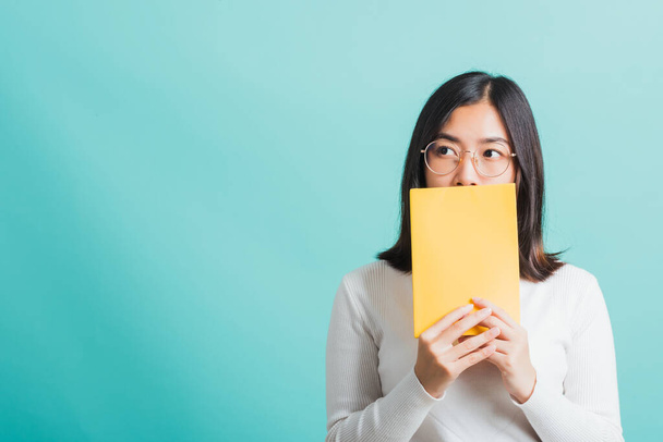 Young beautiful Asian woman hiding behind an open book, Portrait female in glasses is holding and reading a book, studio shot isolated on a blue background, Education concept - Fotoğraf, Görsel