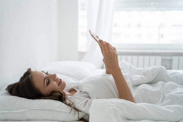 Mujer joven sonriente utilizando el teléfono inteligente móvil, acostado y relajante en la cama cómoda blanca en casa - Foto, imagen