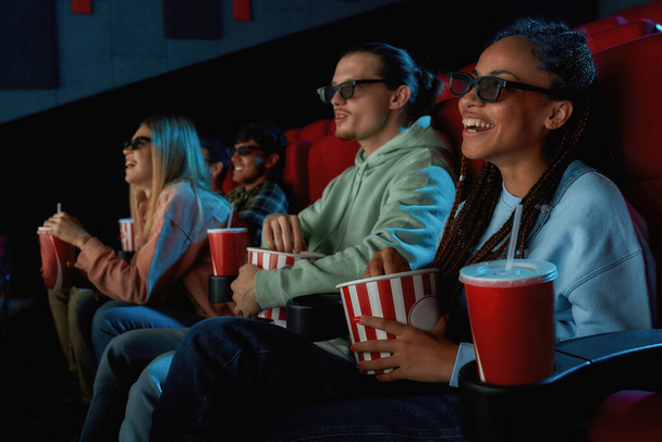 Joyful mixed race young woman wearing glasses, eating popcorn while watching movie in cinema auditorium together with her boyfriend - Foto, afbeelding
