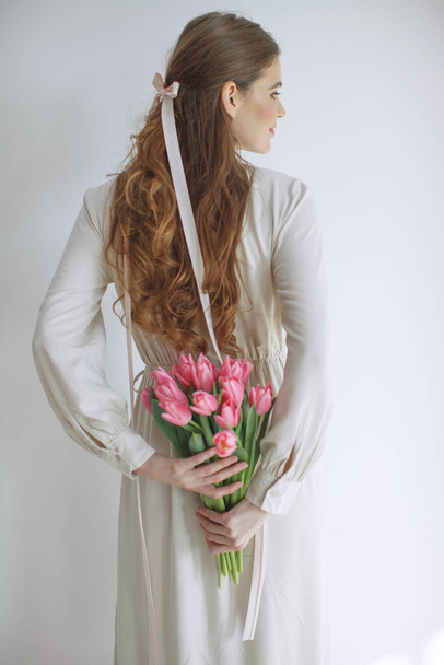 Приваблива молода жінка в сукні з натуральної тканини, що тримає букет тюльпанів, ізольована на білому тлі
. - Фото, зображення
