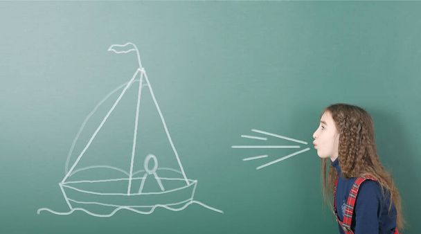 Pre-adolescent girl blowing on painted sailing ship. Portrait photo on school board background. High resolution photo. Full depth of field. - Photo, Image