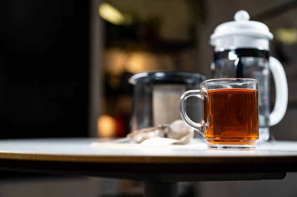 Γυάλινη κούπα φλιτζάνι φρέσκο ζεστό νόστιμο μαύρο τσάι Κεϋλάνης με γαλλικό Τύπο, τσαγιέρα, τσαγιέρα, και το τσάι φίλτρα σε ένα λευκό τραπέζι με από την εστίαση θολή φόντο και πλούσια σκούρα χρώματα - Φωτογραφία, εικόνα