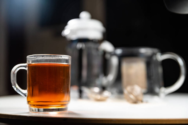 Γυάλινη κούπα φλιτζάνι φρέσκο ζεστό νόστιμο μαύρο τσάι Κεϋλάνης με γαλλικό Τύπο, τσαγιέρα, τσαγιέρα, και το τσάι φίλτρα σε ένα λευκό τραπέζι με από την εστίαση θολή φόντο και πλούσια σκούρα χρώματα - Φωτογραφία, εικόνα