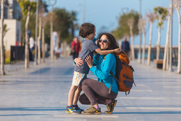 Το αγόρι αγκαλιάζει τη μητέρα του, το παιδί περπατά με τη μητέρα του στο δρόμο της πόλης, η γυναίκα αγκαλιάζει το γιο της. - Φωτογραφία, εικόνα