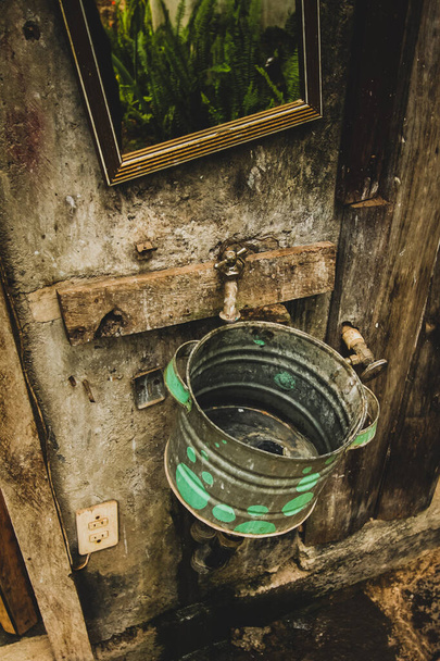 Un viejo cubo de metal envejecido usado como fregadero con un grifo oxidado y un espejo en la pared en Guatemala. - Foto, imagen