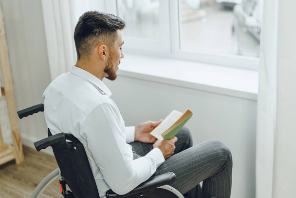 Людина з обмеженими можливостями в інвалідному візку читає книгу в приміщенні
 - Фото, зображення