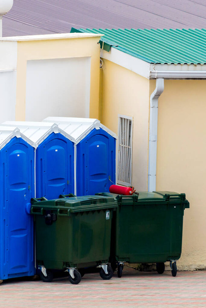 Πράσινοι κάδοι σκουπιδιών με ρόδες και φορητές καμπίνες τουαλέτας σε μπλε. - Φωτογραφία, εικόνα