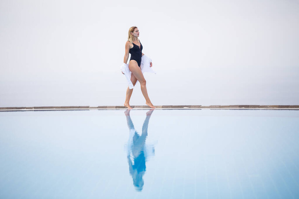 An attractive young woman in a black swimsuit and white shirt stands near the pool with a panoramic view. - Foto, Imagem