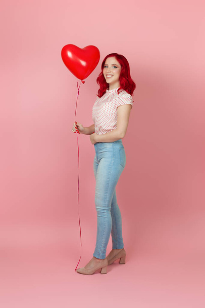 A full-length charming, positive woman with red hair and jeans holds a flying red balloon in her hand , isolated on a pink background - Fotografie, Obrázek