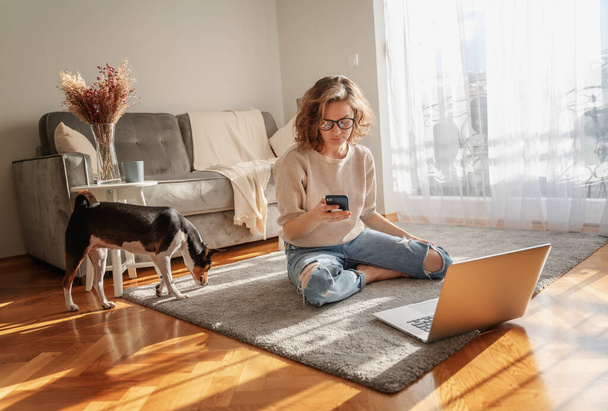 Pretty curly happy young woman sitting at home on the floor in front of laptop with her pet dog working and learning online - Фото, изображение
