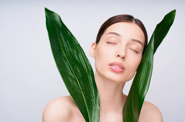 Young pretty sweet gentle girl with clean and fresh skin, dreamily closed her eyes, with green tropical leaves on her face, isolated on white background. Bio, green and ecological cosmetics. - Fotografie, Obrázek