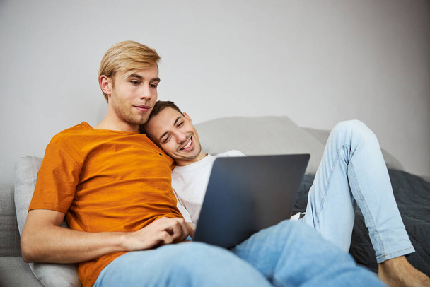 gelukkig gay paar liggend op bank en met behulp van laptop thuis - Foto, afbeelding