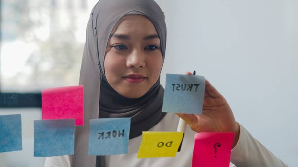 Asiatisch muslimische Dame schreiben Informationen kleben einen klebrigen Zettel auf Glastafel in neuen normalen Büro. Arbeiten von zu Hause aus, Arbeit aus der Ferne, Selbstisolierung, soziale Distanzierung, Quarantäne zur Coronavirus-Prävention. - Foto, Bild