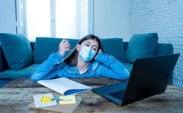 Bored and depressed teenager girl with face mask on laptop studying at home in online education class as high school remain closed due to New COVID-19 lockdown or forecast weather conditions. - Foto, Imagen