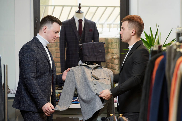 Młody mężczyzna w białym swetrze i garniturze konsultuje się ze sprzedawcą w sprawie koszuli w sklepie odzieżowym. Mężczyzna klient i sprzedawca w butiku. - Zdjęcie, obraz