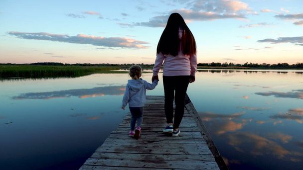 Maman et petite fille marchent sur le pont au coucher du soleil. Il y a un lac autour du pont. Nuages reflétés dans le lac - Photo, image