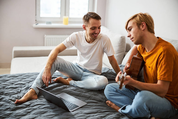 Joyful young man watching how his boyfriend playing guitar - Fotoğraf, Görsel