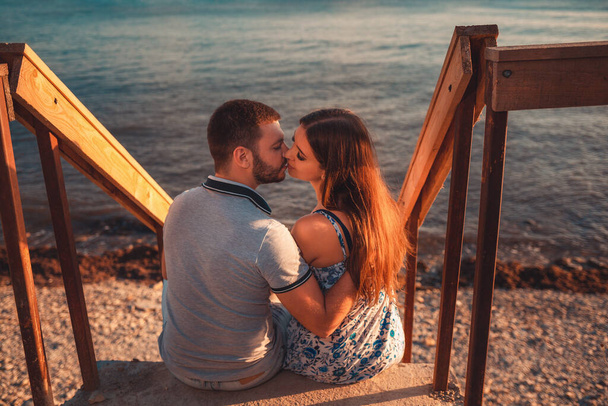 Портрет пары влюбленных обнимает сидящих на лестнице и позирует на фоне моря. Вид сзади. Концепция Дня Святого Валентина. - Фото, изображение