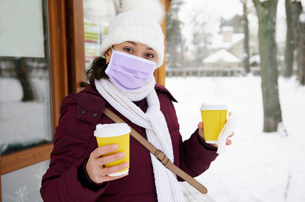 Brünette Frau mit medizinischer Maske mit zwei gelben Tassen Kaffee oder Tee zum Mitnehmen - Foto, Bild