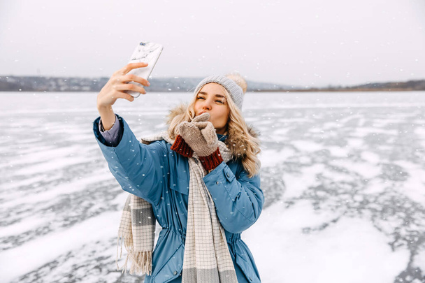 Весела молода блондинка має відеодзвінок, стоячи на замерзлому озері взимку
. - Фото, зображення