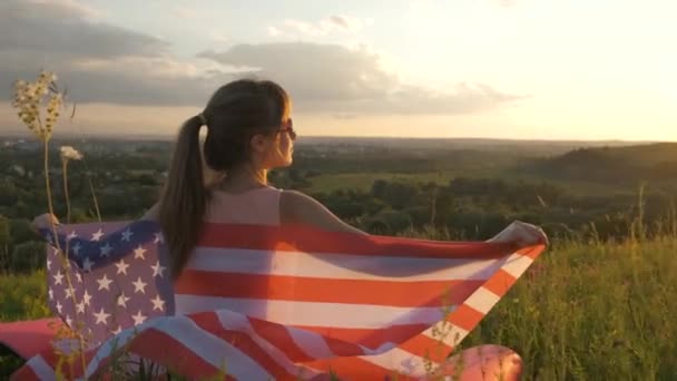 Vissza kilátás boldog fiatal nő fut az USA nemzeti zászló áll a szabadban naplementekor. Pozitív lány ünnepli az Egyesült Államok függetlenségének napját. A demokrácia nemzetközi napja koncepció. - Felvétel, videó