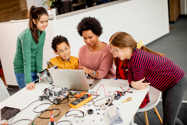 アフリカ系アメリカ人女性科学教師とロボット教室で電気玩具やロボットをラップトップでプログラミングしている幸せな子供たちのグループ - 写真・画像