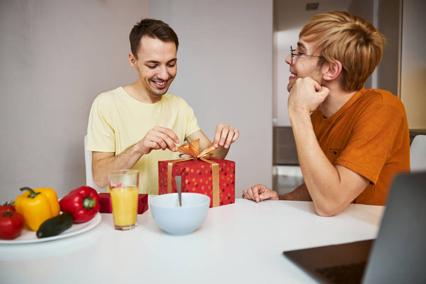 Cheerful guy sitting at the table with boyfriend and opening present - Fotografie, Obrázek