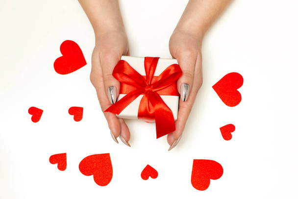 Dámské ruce drží dárek s červenou mašlí na bílém pozadí s papírovými červenými srdci. Backdrop pro: Narozeniny, Valentýn, Vánoce, Nový rok. Plocha - Fotografie, Obrázek