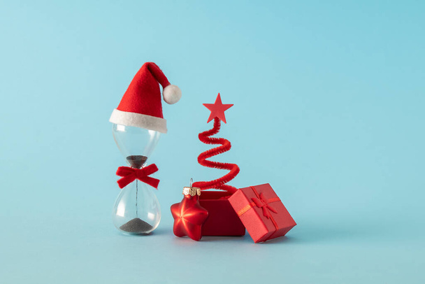 Hourglass with santa claus hat and surprise box with Christmas tree. New year is coming. Holiday creative concept. - Photo, Image