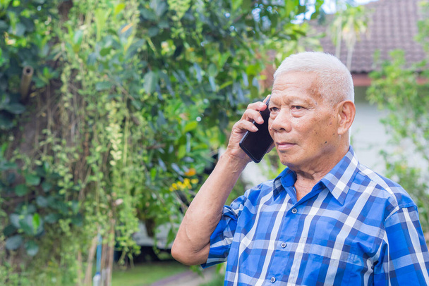 Portré egy idős ázsiai férfiról, aki okostelefont használ, miközben a kertben áll. Helyet a szövegnek. Az idősek fogalma és a kommunikáció. - Fotó, kép