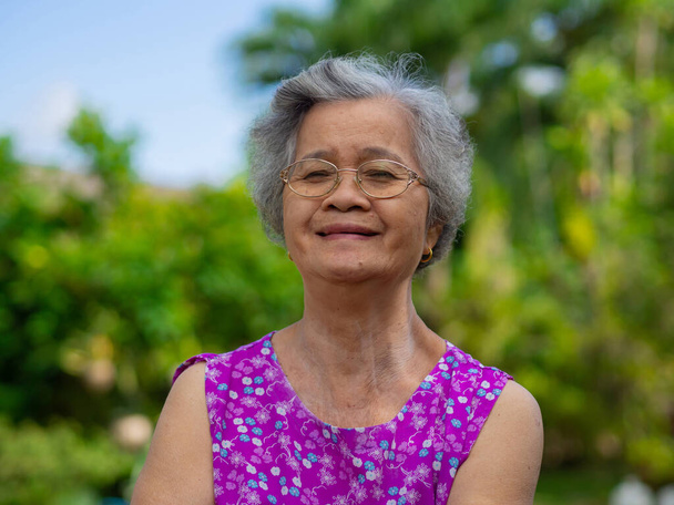 Старшая женщина в очках, улыбается и смотрит в камеру, стоя в саду. Пространство для текста. Концепция пожилых людей и здравоохранения. - Фото, изображение