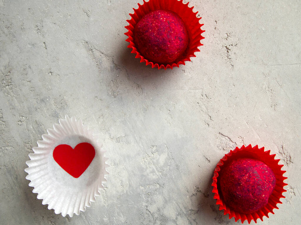 Two appetizing pink candies in red candy wrappers and a red heart in a white wrapper on a gray background. Delicious dark chocolate truffles decorated with sublimated raspberries. - Photo, Image