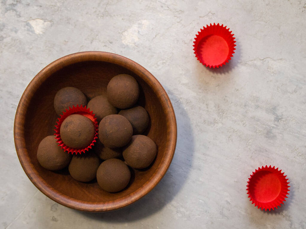 Zwarte chocolade en cacao truffels in een bruin bord. Een van de heerlijke truffels valt op tussen de rest - het is in een rode snoepverpakking. Twee lege snoeppapiertjes liggen naast een kleiplaat. - Foto, afbeelding