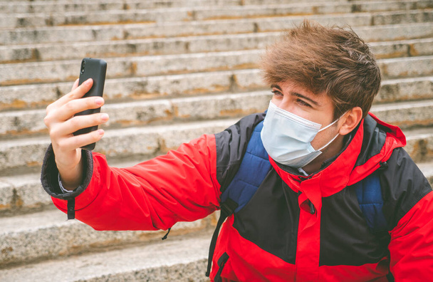 Chlápek s obličejovou maskou a batohem se selfie na schodech na ulici v zimní den. - Fotografie, Obrázek