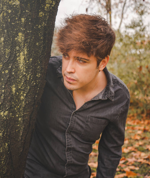 Chlápek, který se opírá o strom a dívá se v zamračeném podzimním dni do lesa. - Fotografie, Obrázek