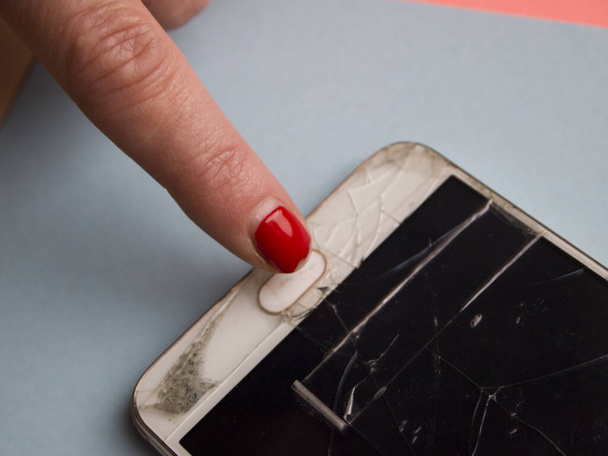 Woman is trying to turn on broken mobile phone. Phone screen is fractured and covered with cracks and sealed with tape. Woman has a sloppy old manicure, which is almost peeled off. Concept of poverty. - Foto, imagen